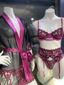 Mannequins displaying a matching lingerie set and robe in a store display. The lingerie set is vibrant magenta with intricate black lace detailing in a heart pattern. The bra has thin magenta straps and sheer lace cups with heart designs, paired with a high-waisted garter belt and matching panties in the same color scheme. Next to it, a black sheer robe with magenta satin trim and heart lace accents complements the set. The robe has a matching satin belt tied at the waist, adding an elegant touch to the display.