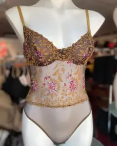 A mannequin wearing a sheer bodysuit in a warm brown color with intricate floral embroidery in gold and pink. The bodysuit has delicate lace detailing along the cups and torso, with thin shoulder straps. The embroidered floral patterns add an elegant, decorative touch to the design.
