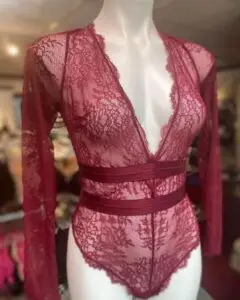 A burgundy lace long-sleeve bodysuit displayed on a mannequin. The bodysuit features a plunging V-neckline, sheer floral lace fabric, and two solid horizontal bands around the waist, adding structure to the delicate lace design.