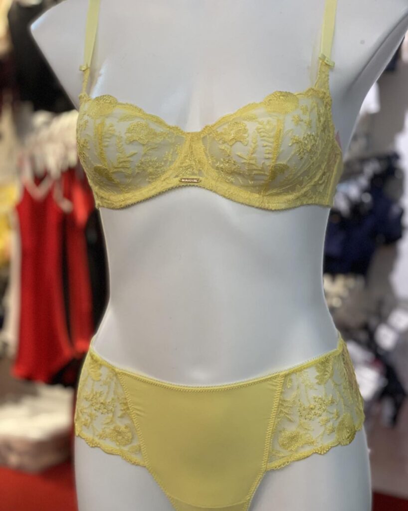 A delicate yellow lingerie set featuring a sheer lace bra with floral embroidery and matching lace-accented panties, displayed on a mannequin in a boutique setting.