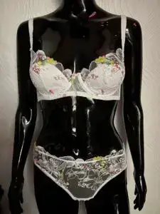 A glossy black mannequin wearing a white lace lingerie set with colorful floral embroidery. The bra has sheer lace cups adorned with pink, yellow, and green floral details, while the matching panties feature similar embroidery with a scalloped lace trim. The mannequin is positioned against a textured white wall background.