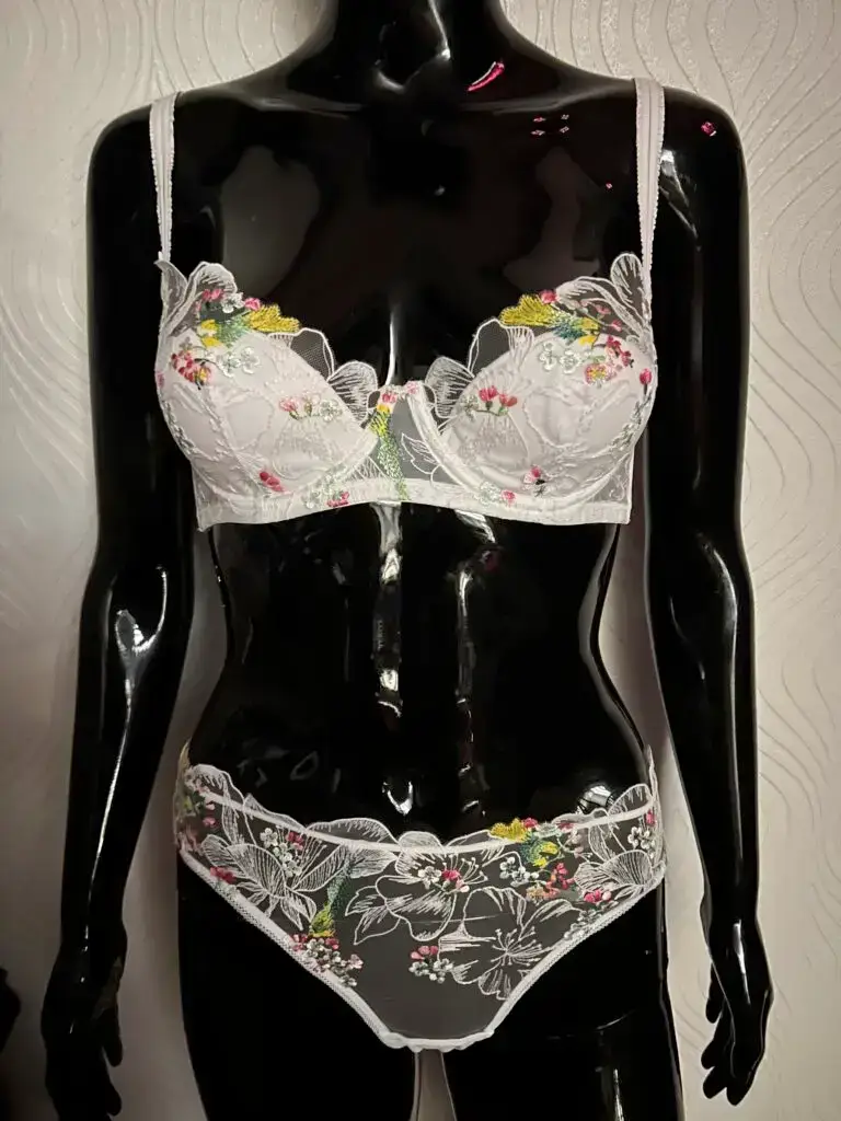 A glossy black mannequin wearing a white lace lingerie set with colorful floral embroidery. The bra has sheer lace cups adorned with pink, yellow, and green floral details, while the matching panties feature similar embroidery with a scalloped lace trim. The mannequin is positioned against a textured white wall background.