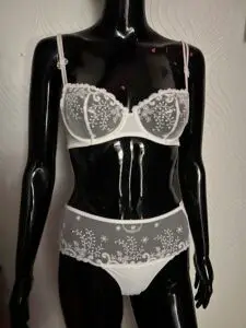 A glossy black mannequin wearing a white lace lingerie set. The bra features intricate sheer lace with floral embroidery and a small satin bow in the center. The matching panties have a combination of lace and opaque fabric with delicate embroidery along the waistband and sides. The mannequin is positioned against a textured white wall background.