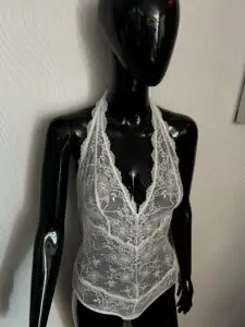 A glossy black mannequin wearing a white lace bodysuit with a halter neckline. The sheer lace fabric features an intricate floral pattern, with scalloped lace trim along the neckline and edges. The mannequin is posed against a textured white wall background.