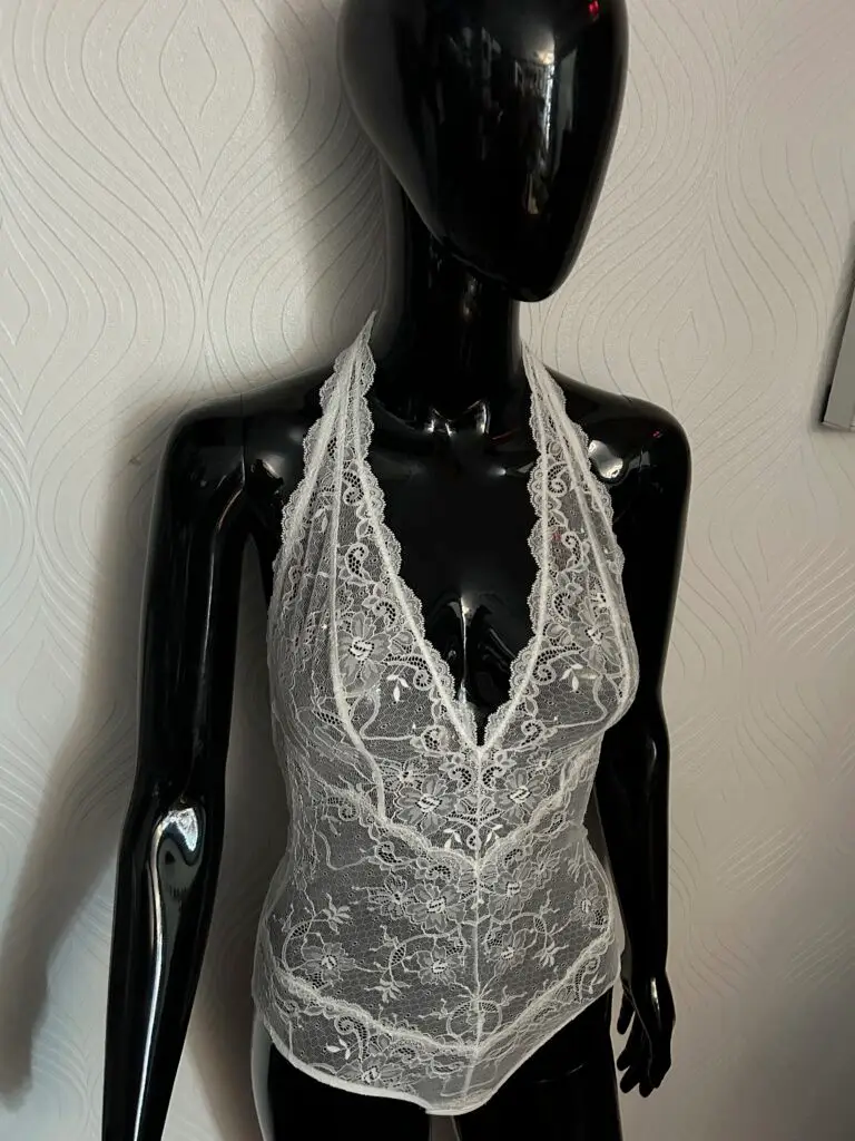 A glossy black mannequin wearing a white lace bodysuit with a halter neckline. The sheer lace fabric features an intricate floral pattern, with scalloped lace trim along the neckline and edges. The mannequin is posed against a textured white wall background.