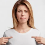 A woman with shoulder-length blonde hair wears a plain white T-shirt and points to her chest with both hands. She has a slightly furrowed brow and a concerned, uncertain expression, suggesting discomfort or dissatisfaction—possibly related to how her bra fits or feels.