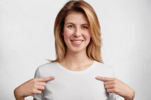 The same woman, now smiling confidently, points to her chest with both hands. Her expression is happy and self-assured, suggesting she feels supported and comfortable—likely due to wearing a properly fitting bra.