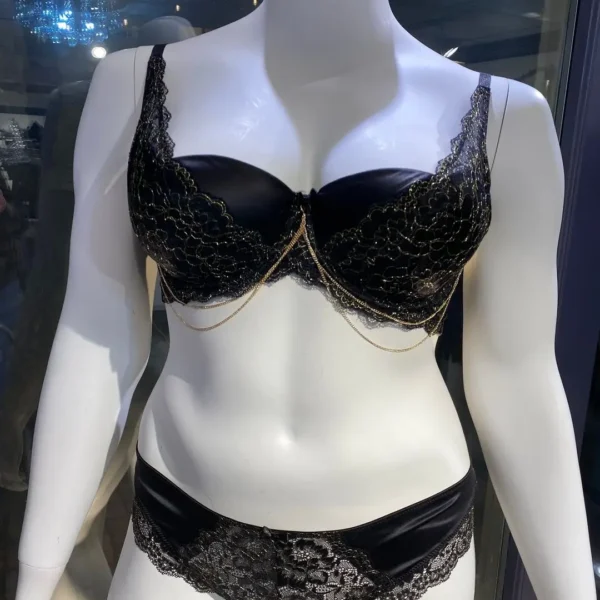 A black bra and panty lingerie set displayed on a mannequin. The bra features lace detailing with delicate gold chain accents draping along the cups, while the matching panties have floral lace across the front, adding elegance to the satin fabric.